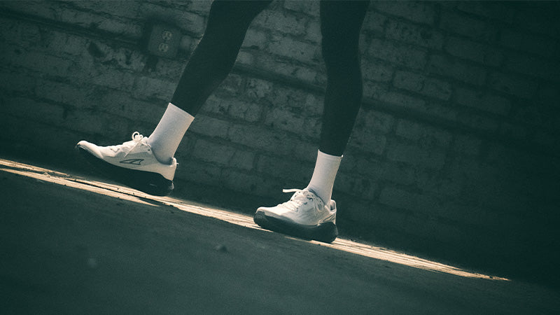 Person wearing white Altra FWD Via running shoes and dark leggings walking on a sidewalk with a brick wall in the background.
