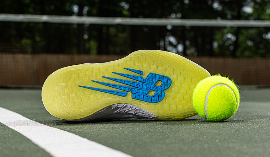 new balance tennis shoe on tennis court with tennis ball, outsole on shoe is showing