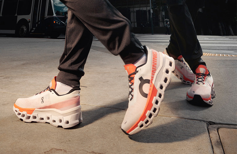 Lifestyle image: closeup of two people walking in On Cloudmonster running shoes on a city sidewalk.