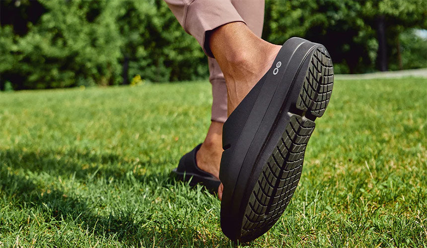 person wearing oofos oomega ooahh sandals in a grassy fieldwalking