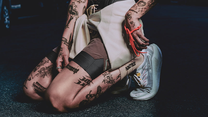 A kneeling person with tattoos wearing Salomon Aero Glide GRVL GTX running shoes