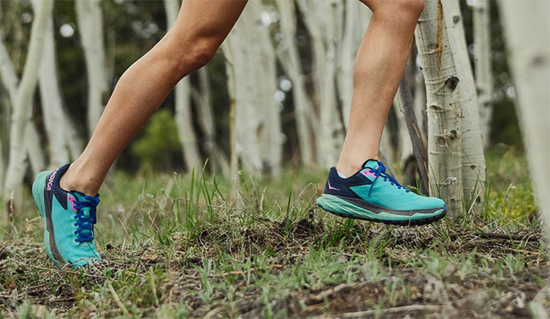 Woman running through green grass and white trees in teal colored Hoka Zinal Atlantis/Outer Space trail running shoes. 