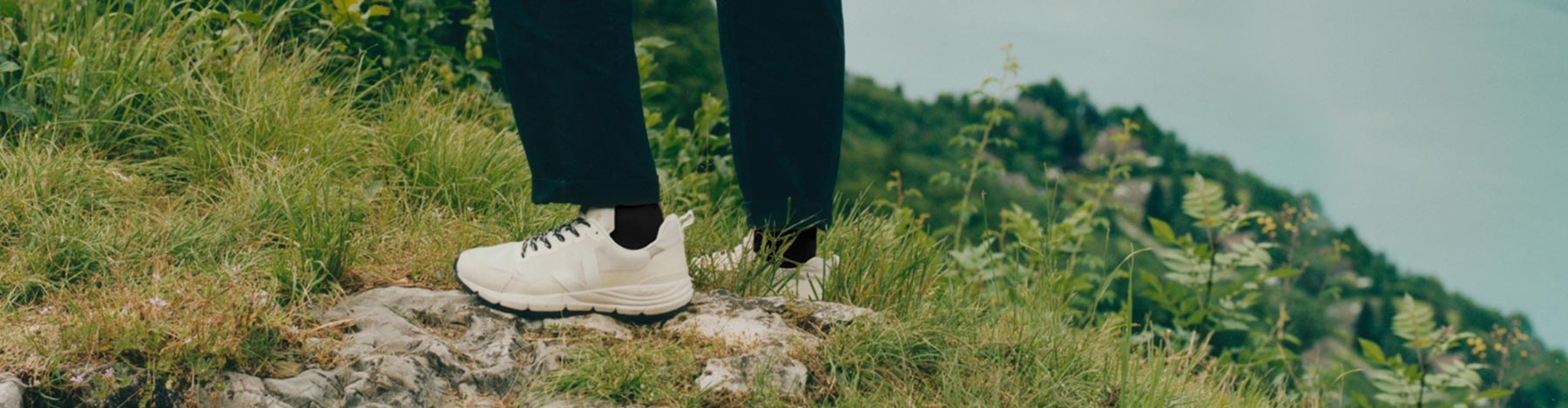 Person standing on a rocky hill overlooking a green forest and body of water in Natural/White VEJA Dekkan hiking shoes.