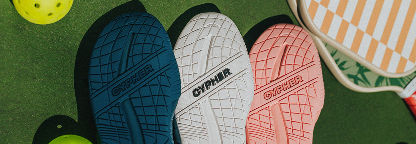 A photo showing the outsoles of the Cypher Pickleball Key211 Pickleball shoes in a variety of colors next to pickleball paddles and balls.