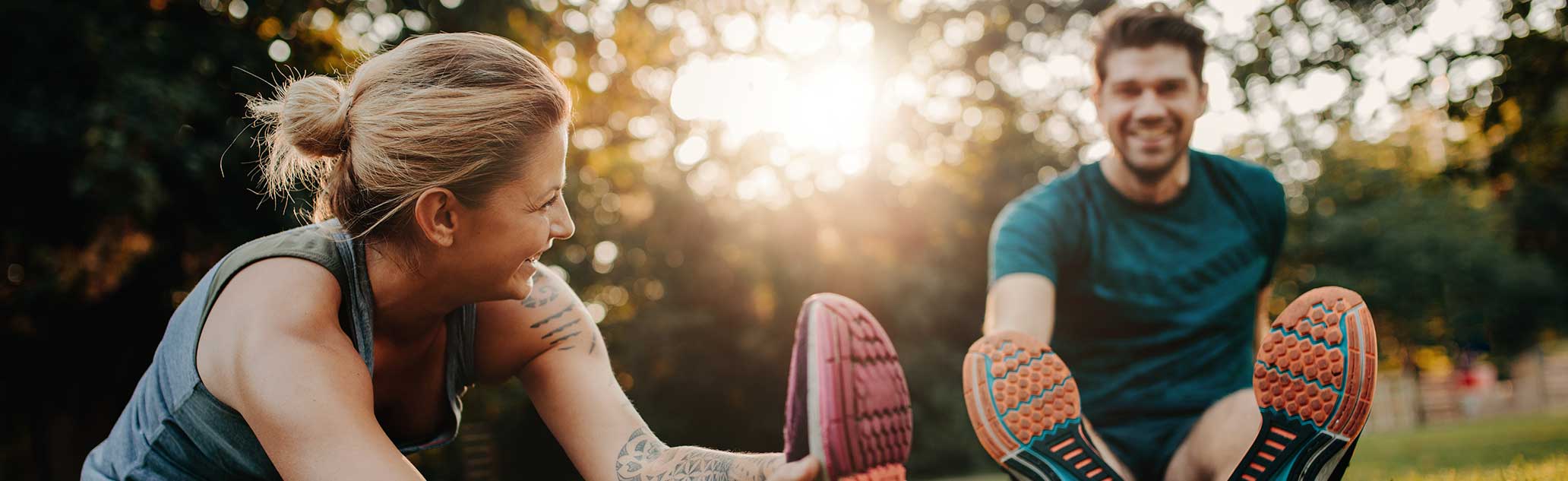 Woman and man seated on grass wearing athletic shoes and smiling at one another.