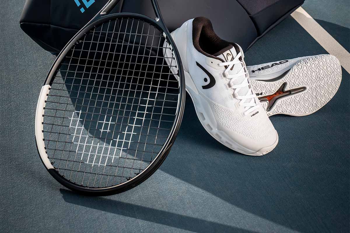 Closeup of a HEAD Speed 2026 tennis racquet and white Revolt Pro 4.5 shoes leaning against a bag on a blue tennis court.