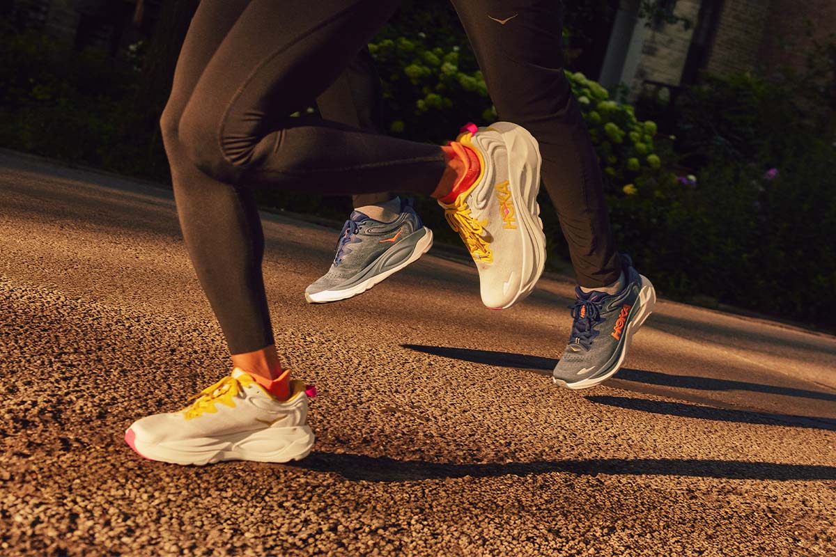 Two runners in motion on a paved path lined with flowers and homes wearing HOKA Gaviota 6 running shoes.