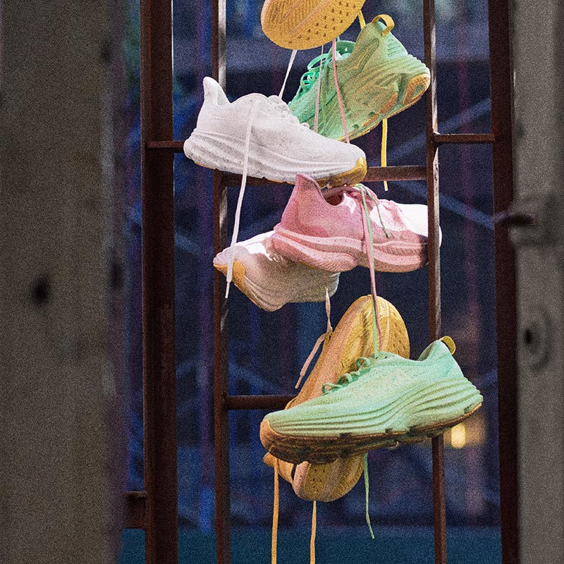 Green, white, pink, and yellow HOKA Global Running Day 2024 shoes hanging from an urban structure.