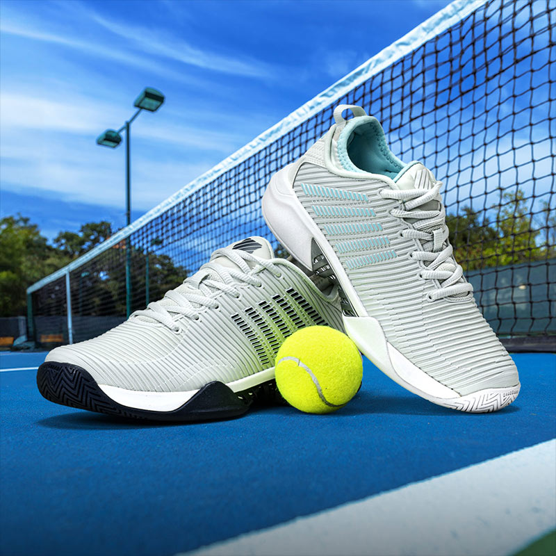 Men's and women's K-Swiss tennis shoes and a tennis ball sitting on a blue tennis court.