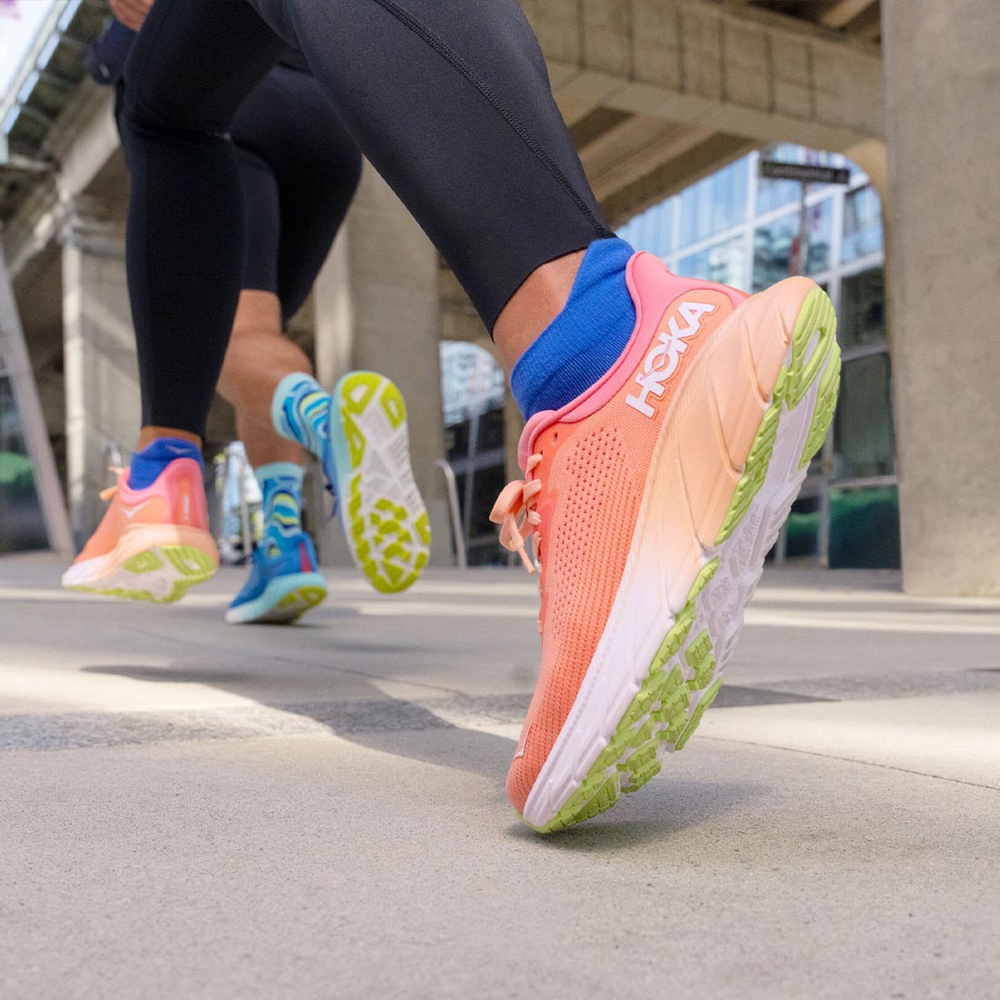 Lifestyle image: Two people running in women's and men's HOKA Arahi 7 running shoes on a city sidewalk.