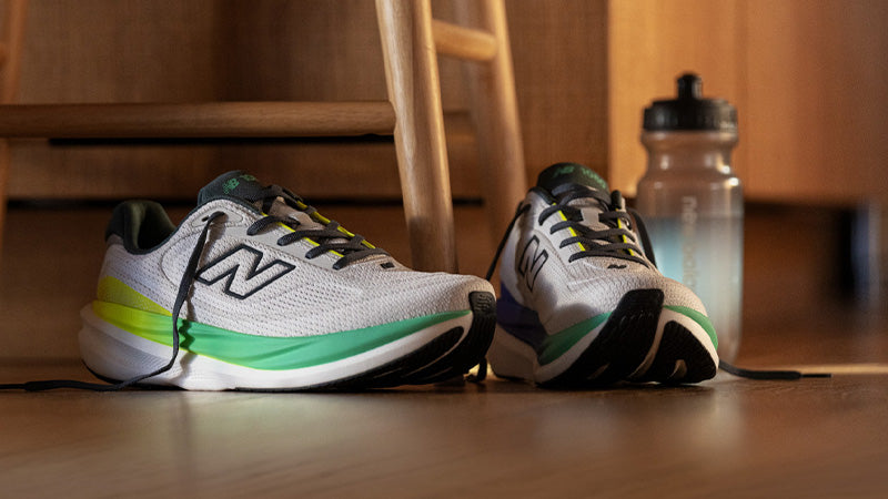 Pair of white New Balance 1080v15 running shoes with green and black accents on a wooden floor next to a wooden stool and a water bottle.