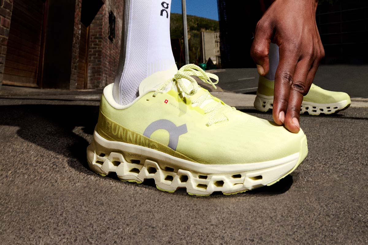 A person stretching in bright yellow On Cloudmonster 3 running shoes on a sidewalk next to a brick building