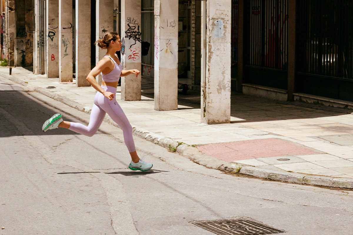 A woman jogging in light lavender On clothing and green On shoes on a city street with stone columns and pillars in the background.
