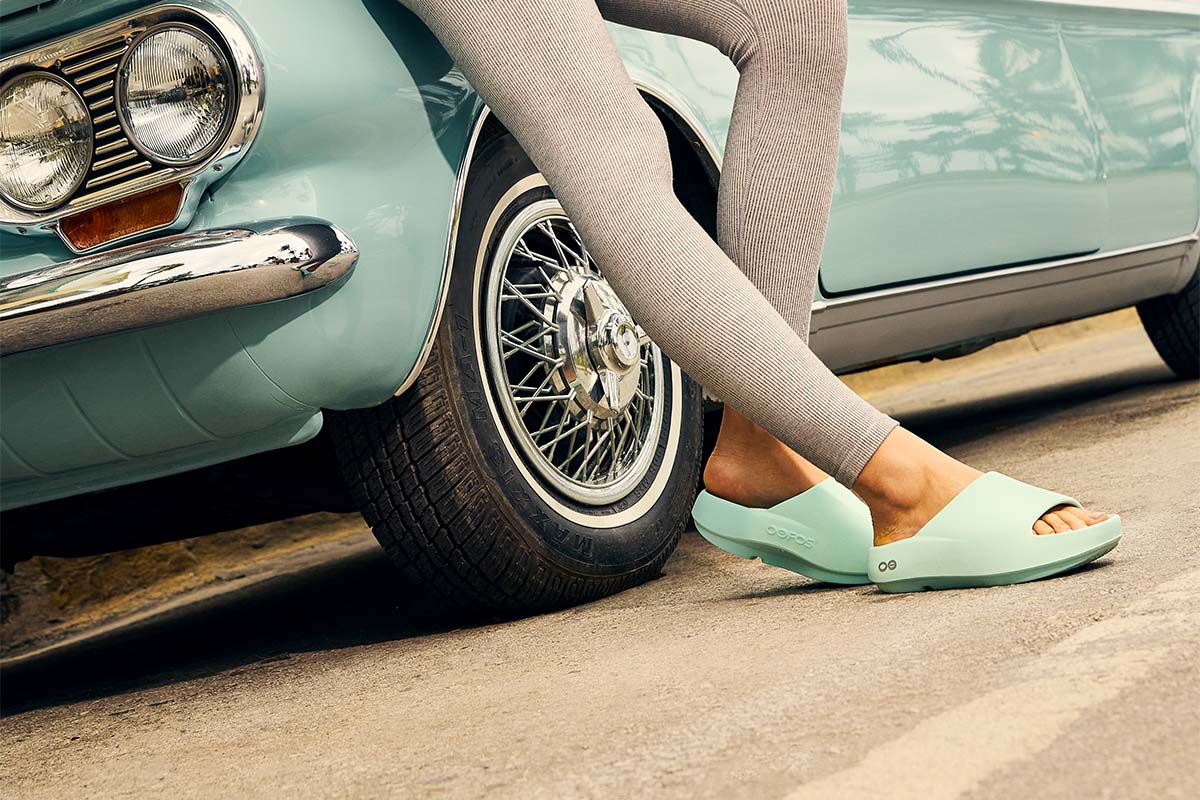 Person wearing light green OOFOS OOahh+ recovery slides standing next to a light green vintage car.