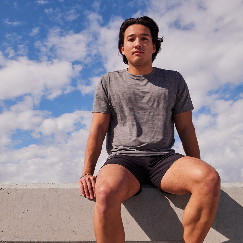 Lifestyle image: a man wearing Brooks running clothing sitting on on a concrete ledge against a blue sky with many white clouds..