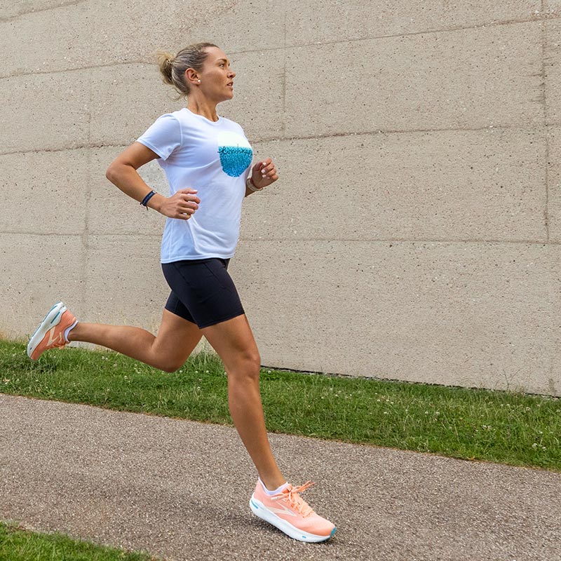 Lifestyle image: a woman in Brooks running clothing and shoes running a path lined with green grass and a concrete wall in the background.