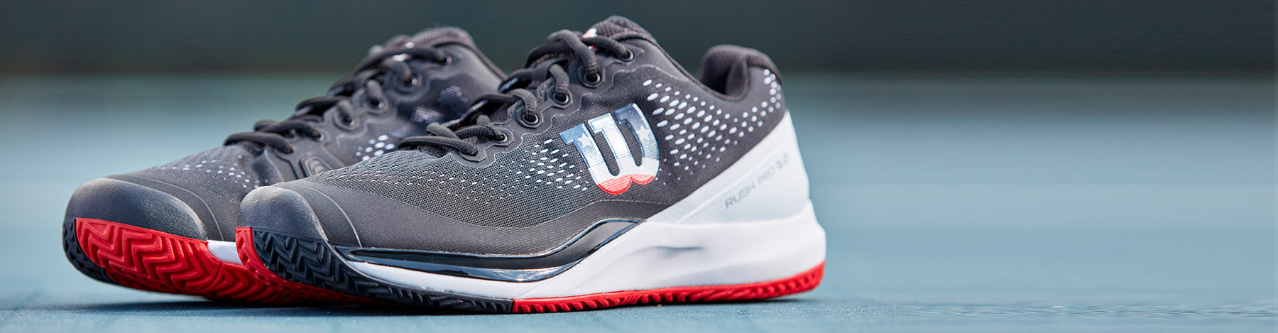 Wilson Rush Pro 3.0 Men's Black/White/Infrared Pickleball Shoes on a light blue outdoor court. 