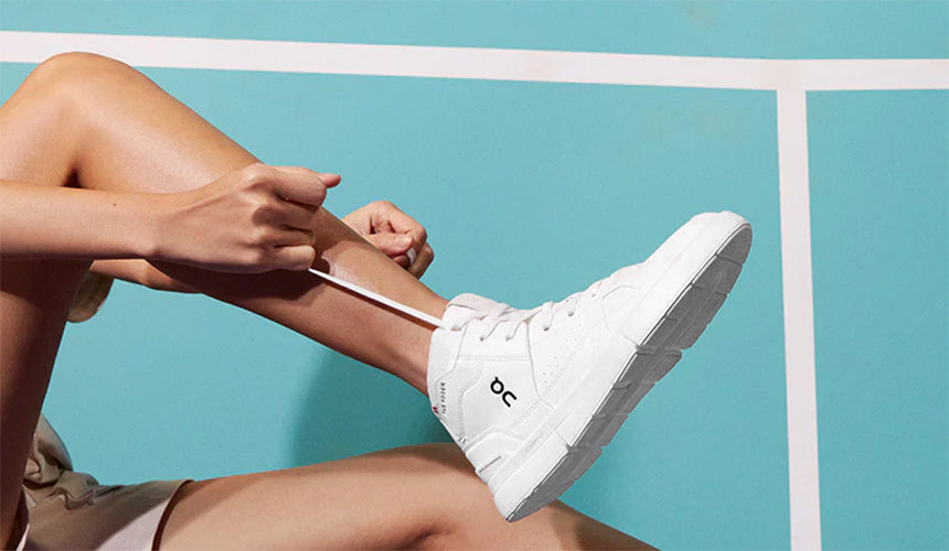A woman lacing up a white pair of On The Roger Clubhouse Mid tennis shoes against a light blue tennis court background.