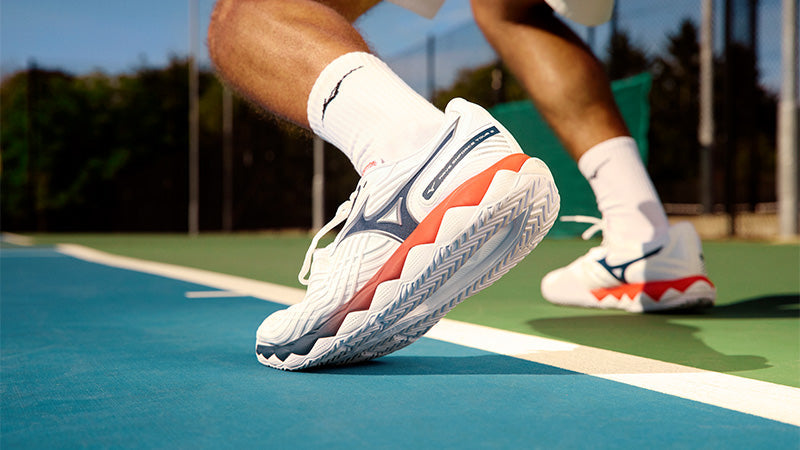 A tennis player on a blue and green court wearing white Mizuno Wave Enforce Tour 2 shoes