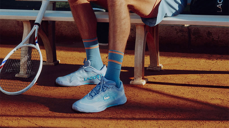 Tennis player sitting on a bench wearing blue Lacoste tennis shoes and socks with a Tecnifibre racquet on a red clay court surface