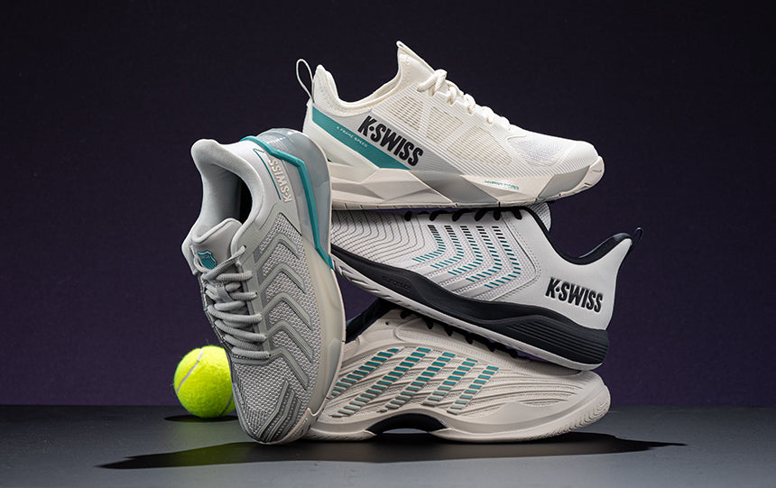 Four K-Swiss tennis shoes stacked on a gray floor against a purple background with a yellow tennis ball.