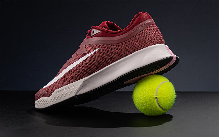 A red Nike Vapor 12 tennis shoe with a tennis ball under the heel on a dark studio background