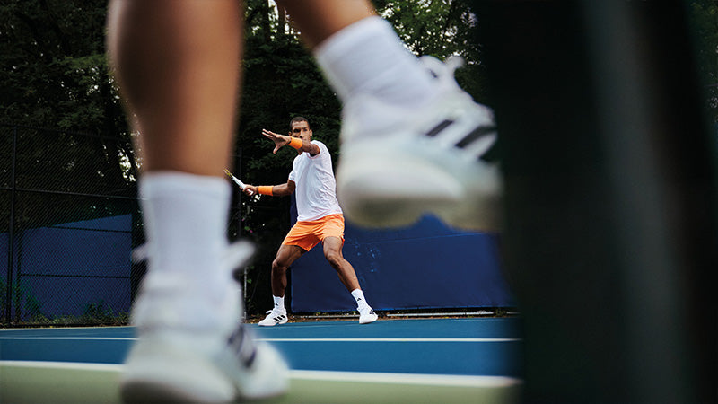 Person playing tennis on a blue court wearing orange and white tennis clothing and shoes with another person's blurred feet and tennis shoes in the foreground