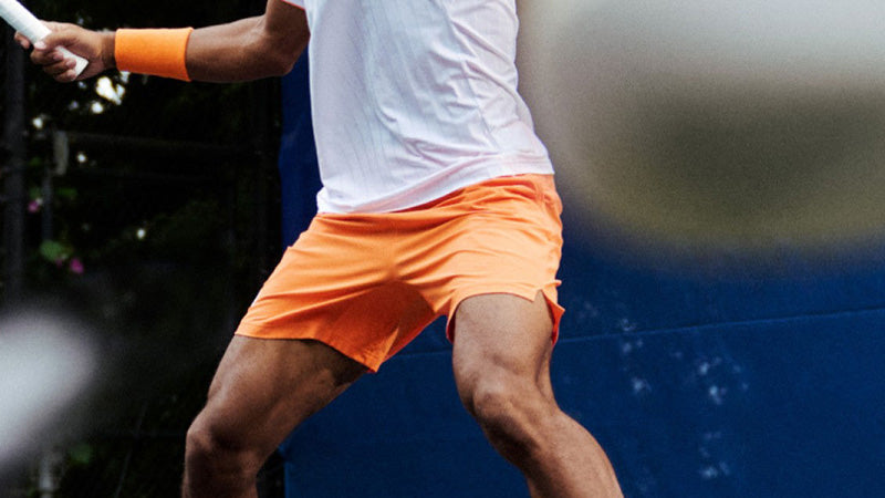 Action shot of a man playing tennis in adidas clothing with the focus on his orange tennis shorts.