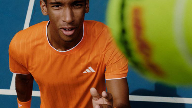 A tennis player wearing an orange adidas tee tossing a tennis ball up at the camera on blue court