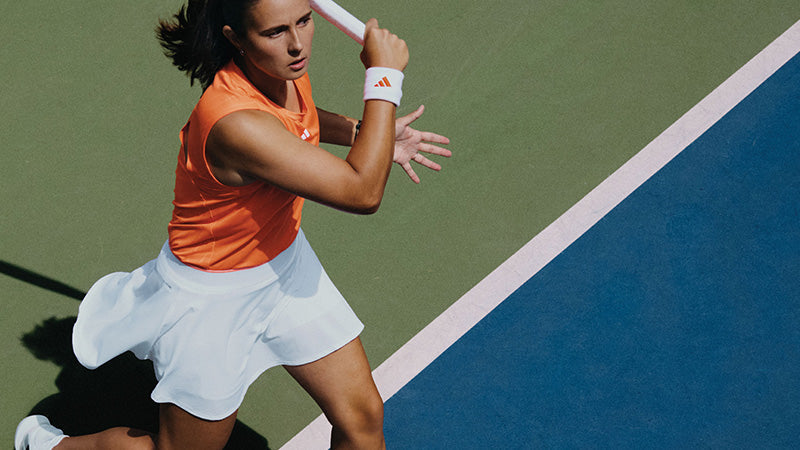 tennis player wearing an orange adidas top and white skirt on a blue and green tennis court