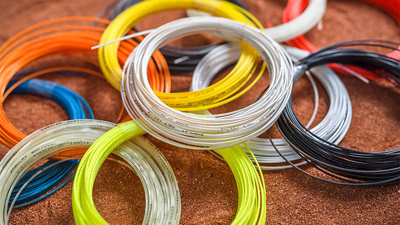 Multiple unpackaged sets of Babolat tennis strings in assorted colors, arranged on a clay tennis court surface