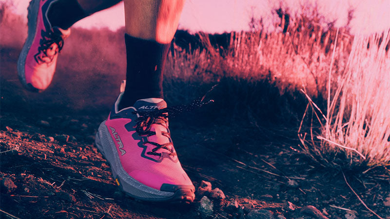 pink-tinted image of a person running a dirt path in pink Altra Experience Wild 3+ trail running shoes with purple accents