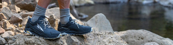A person standing on the rocky edge of a small stream in black Altra Olympus 5 Hike Low GTX Hiking Shoes.