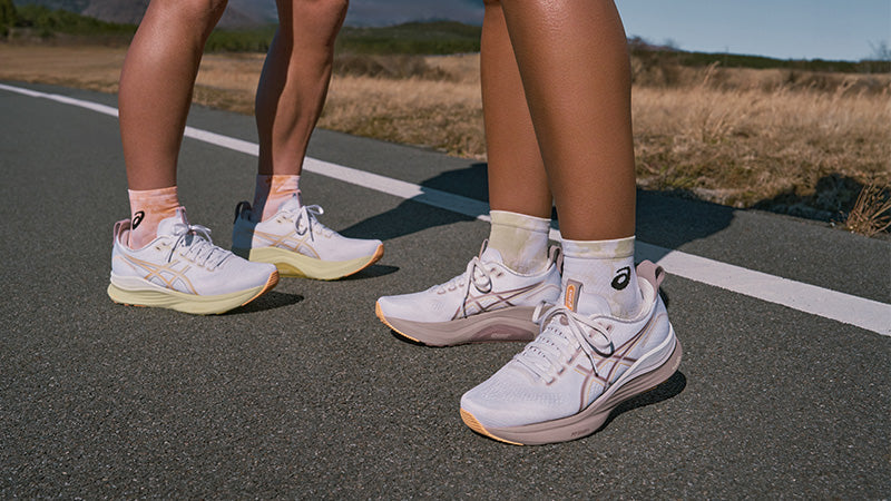 Two people standing on a paved road next to an open field of dry grass wearing white ASICS GEL-Kayano 32 running shoes with orange accents