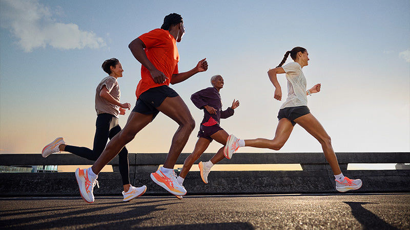 A group of four runners wearing ASICS Rise & Shine pack running shoes on a roadway with a clear sky sunrise in the background.