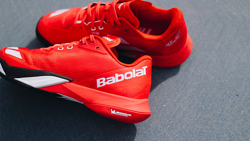 a pair of red Babolat Jet Mach 4 tennis shoes on a blue gray court surface