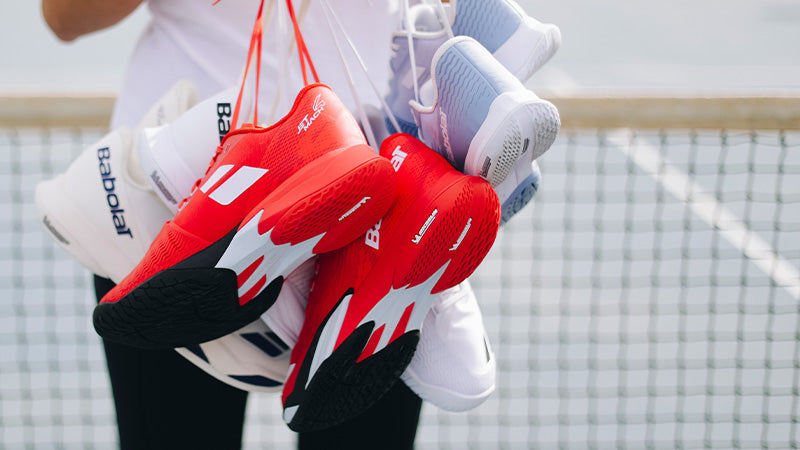 Person holding red, white and powder blue Babolat Jet Mach 4 tennis shoes by the laces, standing in front of a blurred tennis net