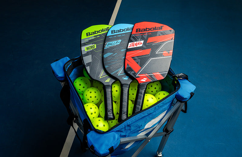 Babolat pickleball paddles and balls sitting in a blue cart on a blue court.