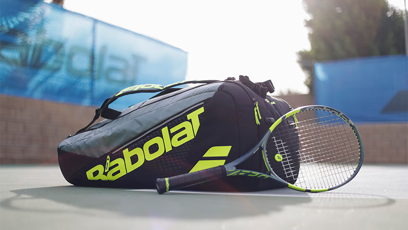 A dark colored Babolat Pure Aero 2026 tennis racquet and bag on a tennis court with neon yellow-green branding and accents.