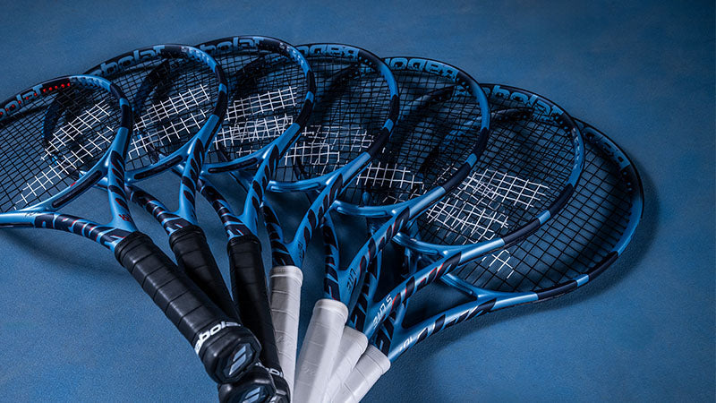 Seven Babolat Pure Drive tennis racquets in blue and black, fanned out on a blue tennis court surface.