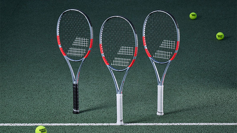 Three Babolat Pure Strike Carbon Grey tennis racquets posed upright on a green tennis court with three tennis balls loosely scattered around them.
