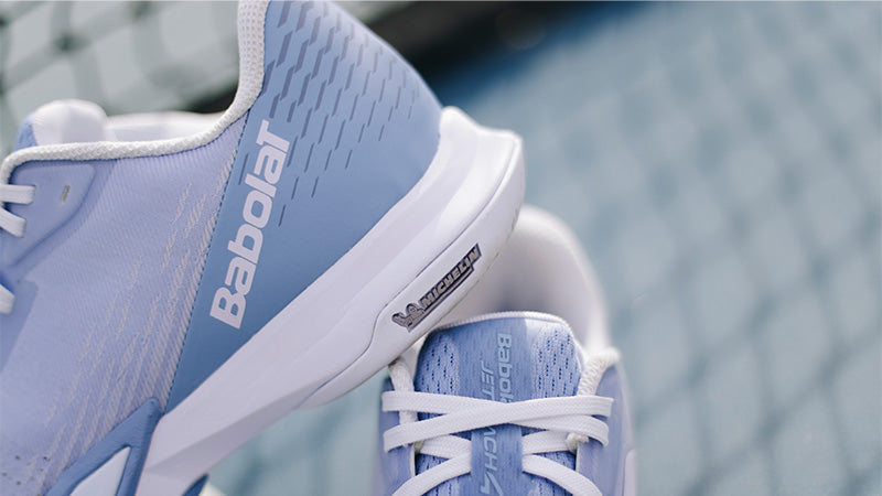 A close-up of a light blue and white Babolat tennis shoe with the brand name 'Babolat' printed on the side, with a blurred tennis court surface and net in the background.