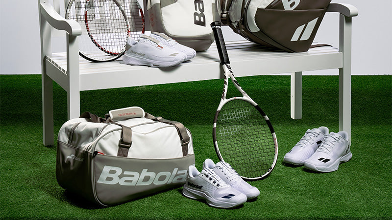 Assorted Babolat Wimbledon tennis shoes, racquets and bags on and around a white bench on a green turf grass surface