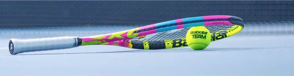 Pink, Blue and Yellow Babolot Pure Aero Rafa 2023 tennis racquet resting on a tennis ball on a blue court.