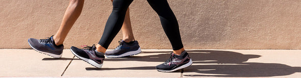 Image of a man and a woman walking on a sidewalk along a tan, textured wall in Brooks Addiction 14 running shoes.