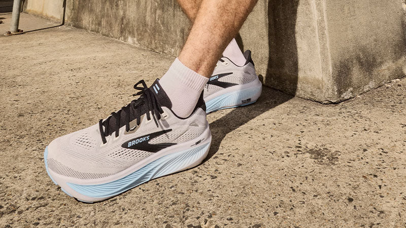 A person wearing light gray Brooks Ghost 18 men's running shoes with light blue and black accents on a concrete surface