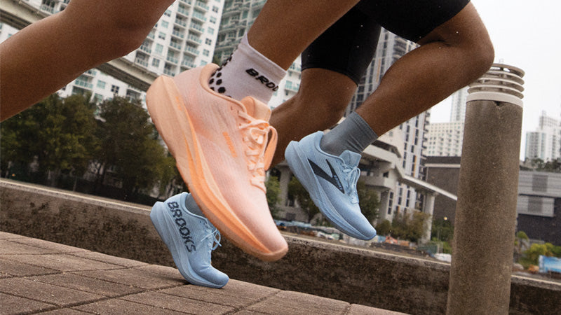 Two runners wearing Brooks Ghost Amp shoes on a concrete path with trees and high rise buildings in the background.