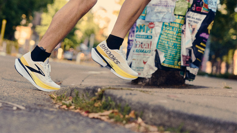 A runner on a city sidewalk wearing Brooks Hyperion Max 4 shoes by a pole with crinkled posters stuck all around it