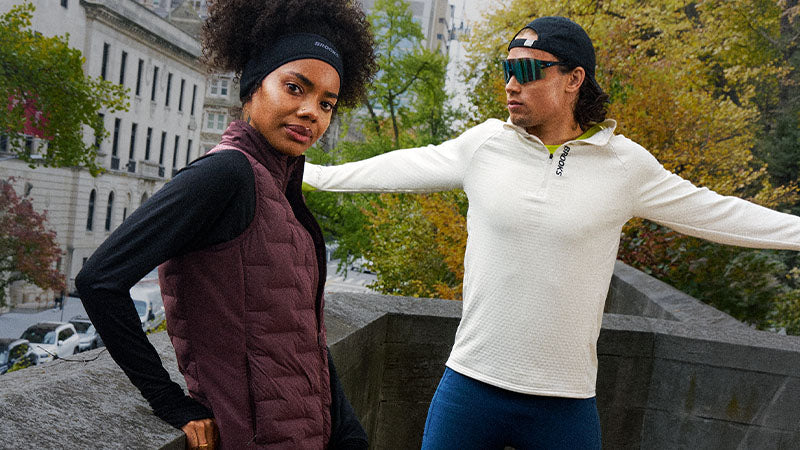 Two people wearing Brooks running clothing and headwear for fall weather, outdoors in a city with trees and buildings in the background