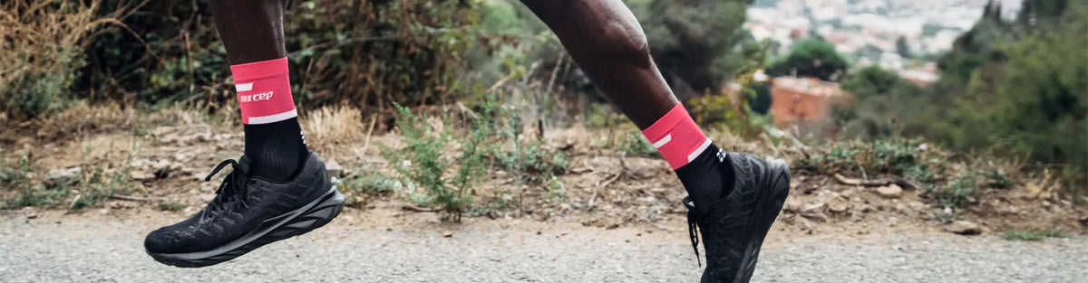 A man running on the side of a hilltop road in pink and black CEP The Run Compresssion Mid Cut Socks 4.0 and black running shoes.